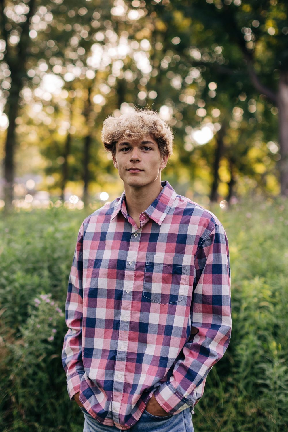 Someone wearing a plaid button-down shirt poses in front of lush greenery during golden hour.