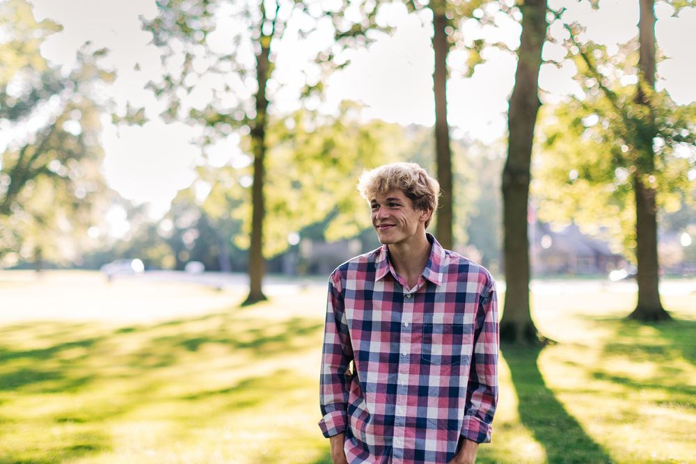 A person wearing a plaid shirt stands in a sunlit park with trees in the background and golden sunlight filtering through.