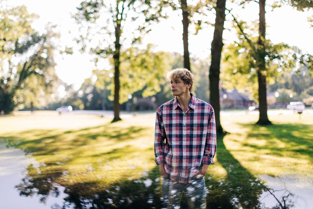 A person in plaid shirt stands outdoors in a park with trees and sunlit grass on a bright autumn day.
