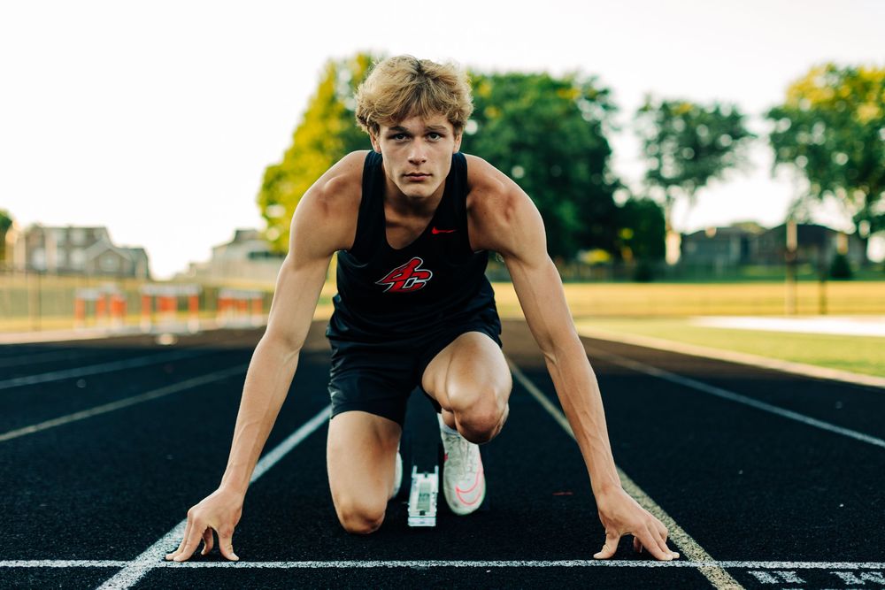 Athlete in dark tank top crouches in starting position on running track during golden hour at sports stadium.