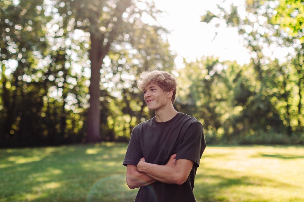 Person in black t-shirt standing with arms crossed in sunny backyard with trees casting warm afternoon light.