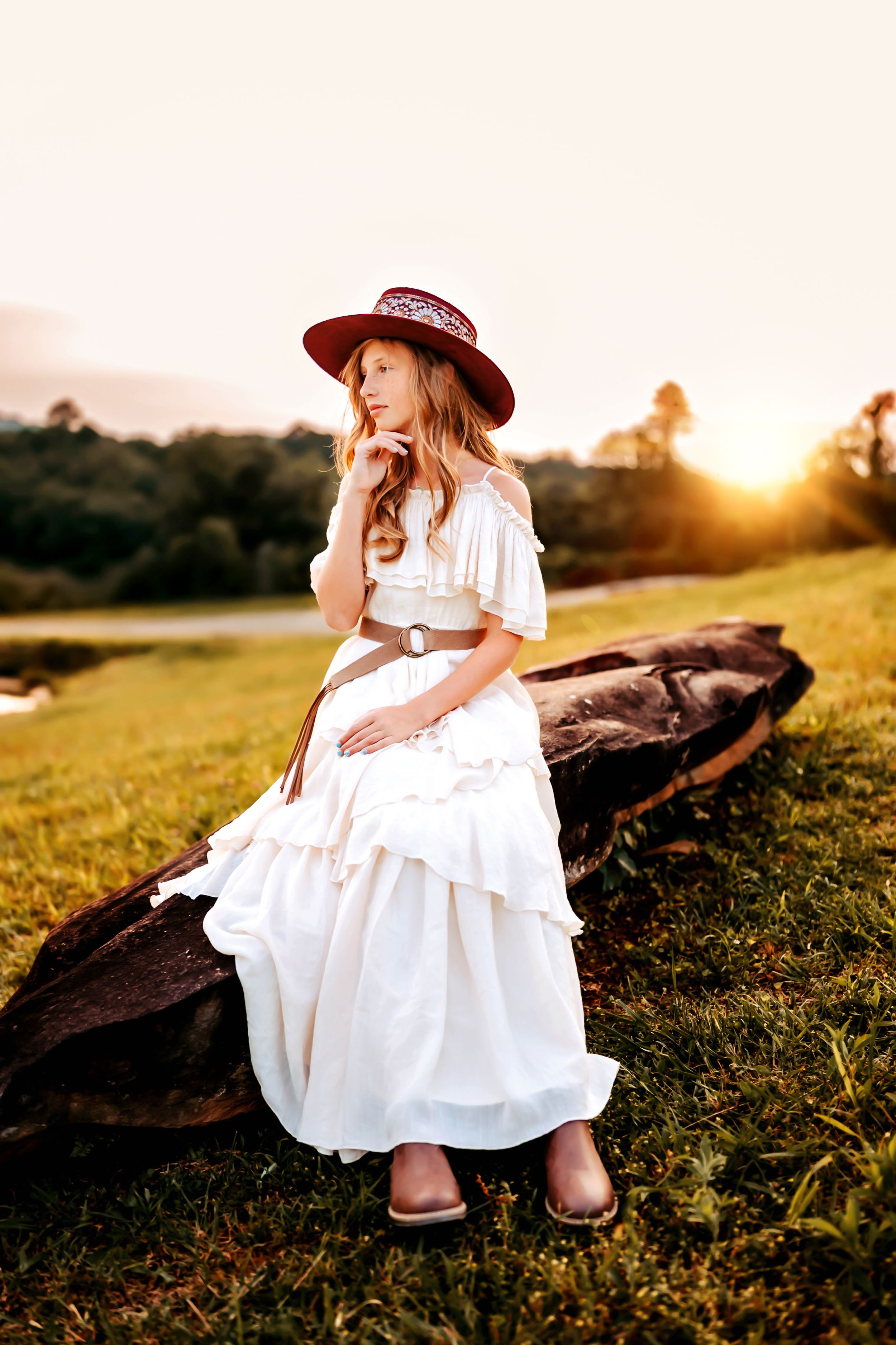 A person in a white flowing dress and burgundy hat sits on rocks in a sunlit meadow during golden hour.