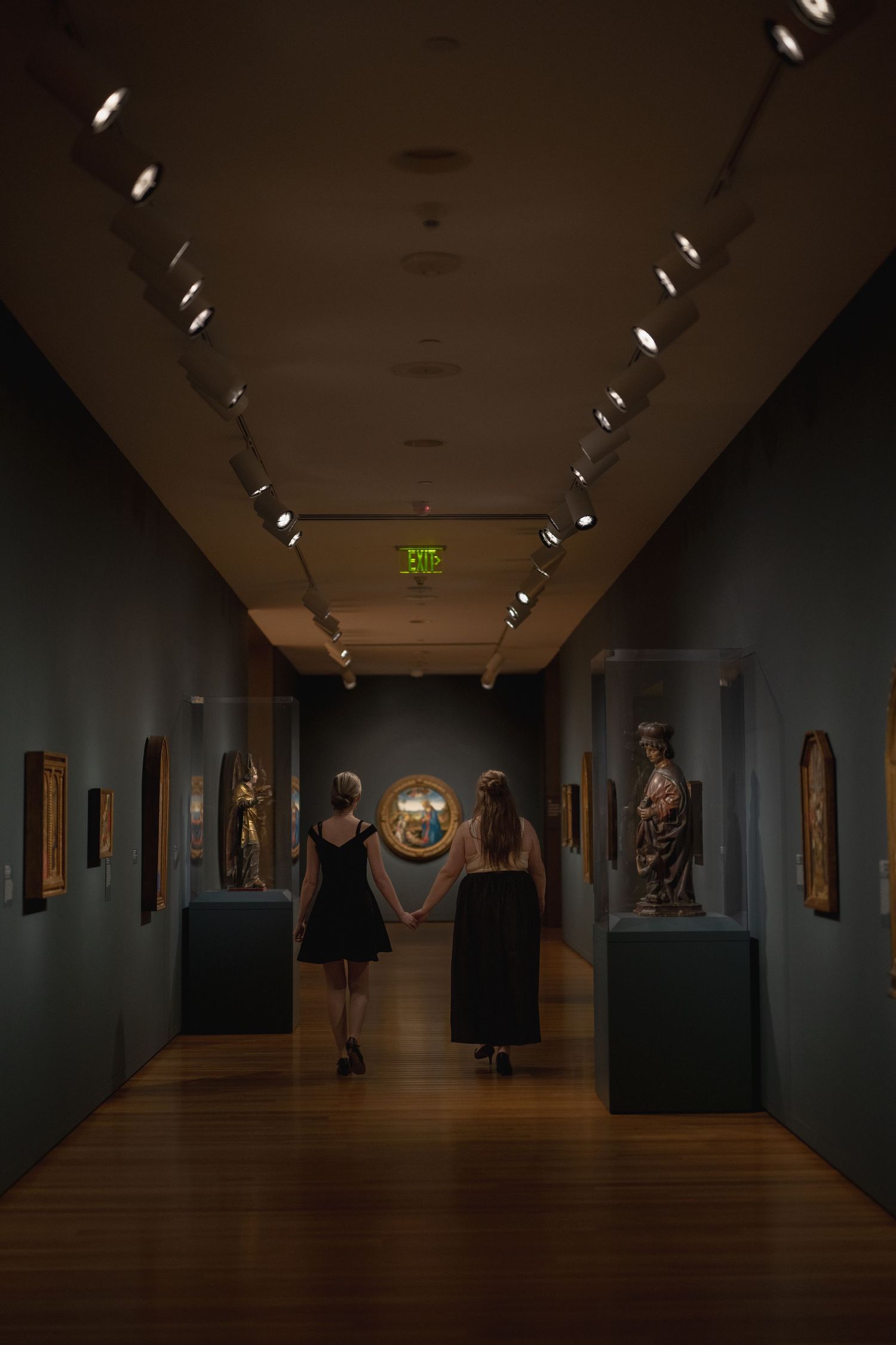 Two figures in black dresses view artwork in a dimly lit museum gallery with spotlights illuminating sculptures and paintings.