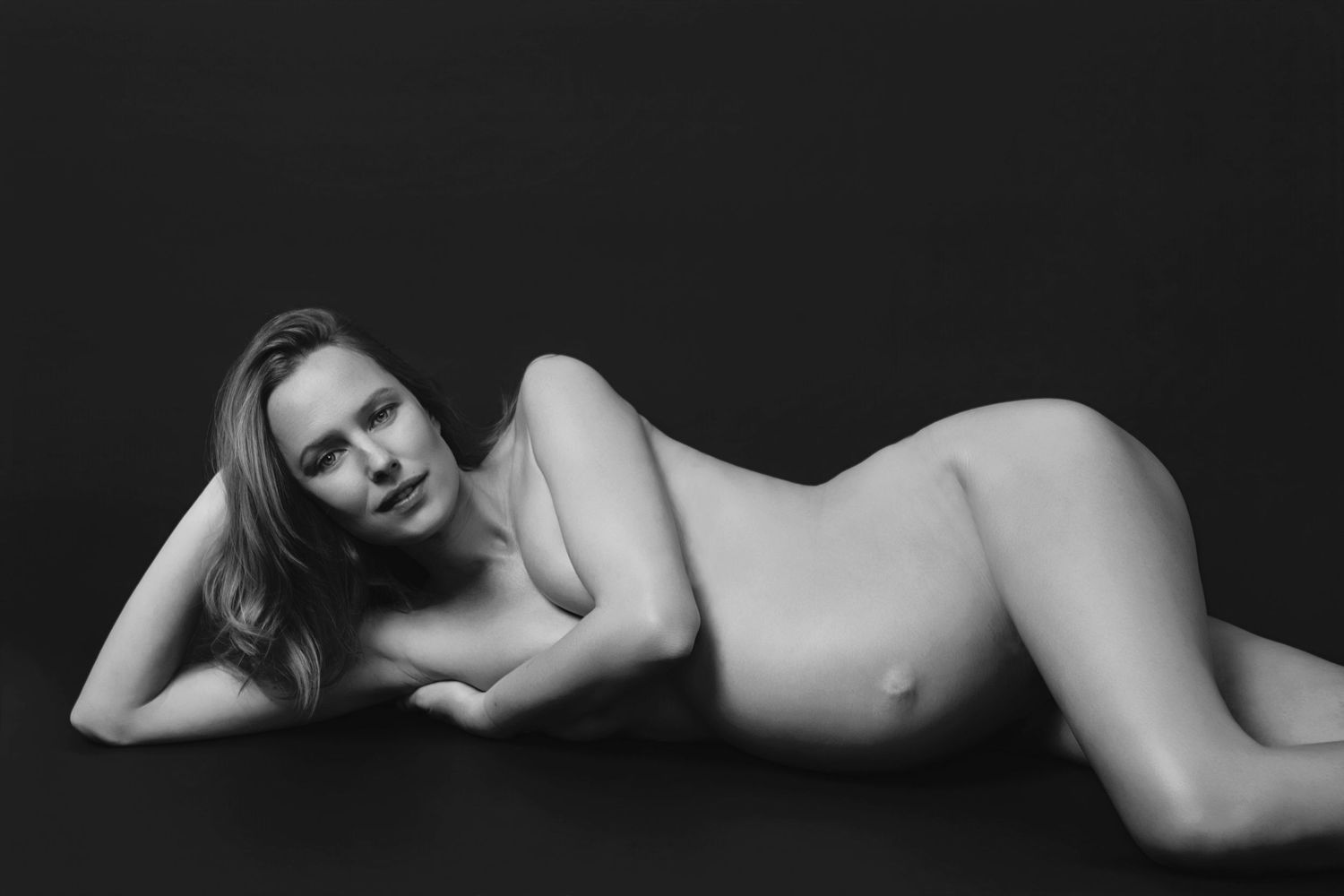 A dramatic low key maternity portrait showing a silhouetted pregnant figure sitting against a dark background.