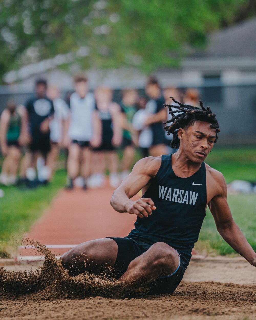 Sports Photography of track athlete in Warsaw singlet performs long jump creating sand spray on landing - Warsaw, Indiana