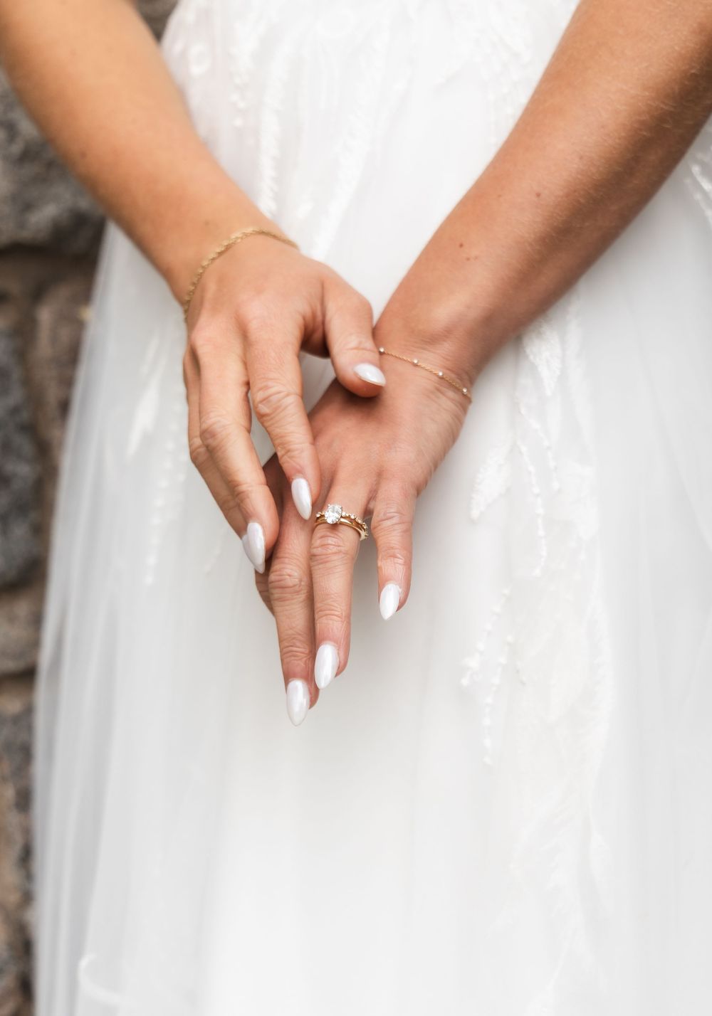 Close-up of bridal hands with manicured nails showing engagement ring against white dress.