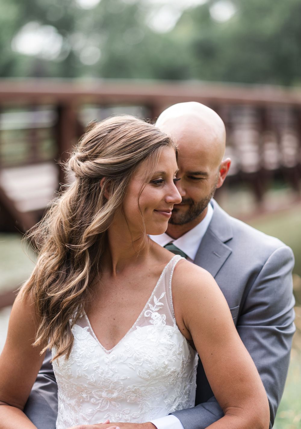 A bride in a lace gown smiles as her bald groom in a gray suit tenderly nuzzles her cheek outdoors.
