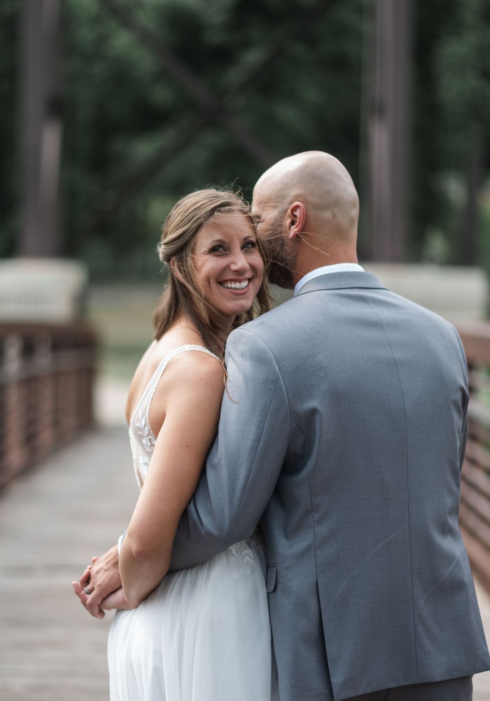 Bride in white dress smiles over shoulder while groom in gray suit embraces her on a wooden bridge.
