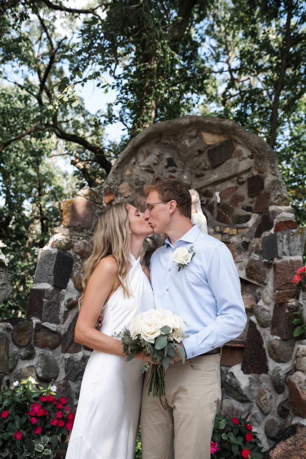 A romantic wedding couple shares kisses in front of a rustic stone wall surrounded by greenery and bright red flowers.