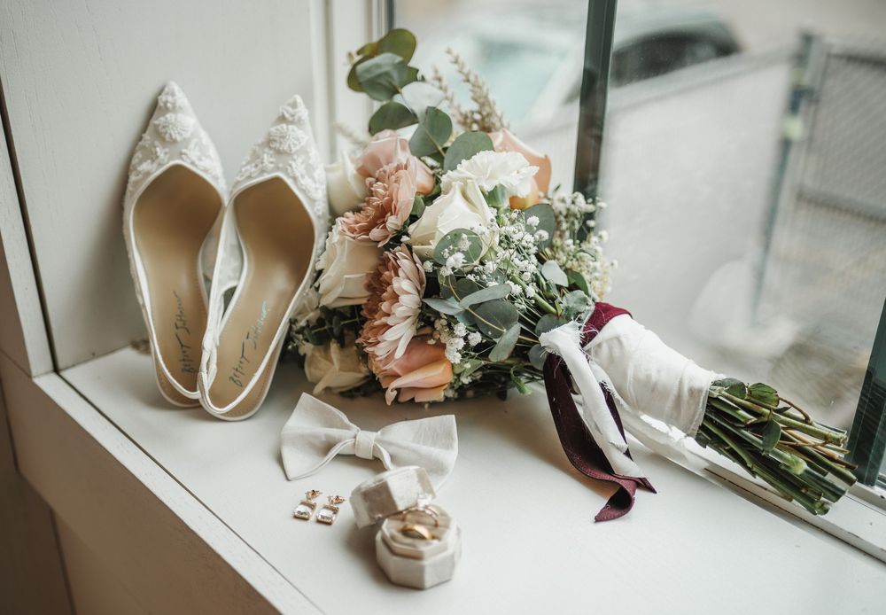 Wedding details including elegant white heels, delicate floral arrangements, and bridal accessories on a windowsill.