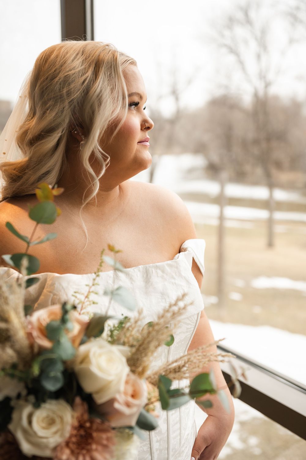 A bride in an off-shoulder white dress holds a romantic bouquet of roses and eucalyptus while gazing out a window.