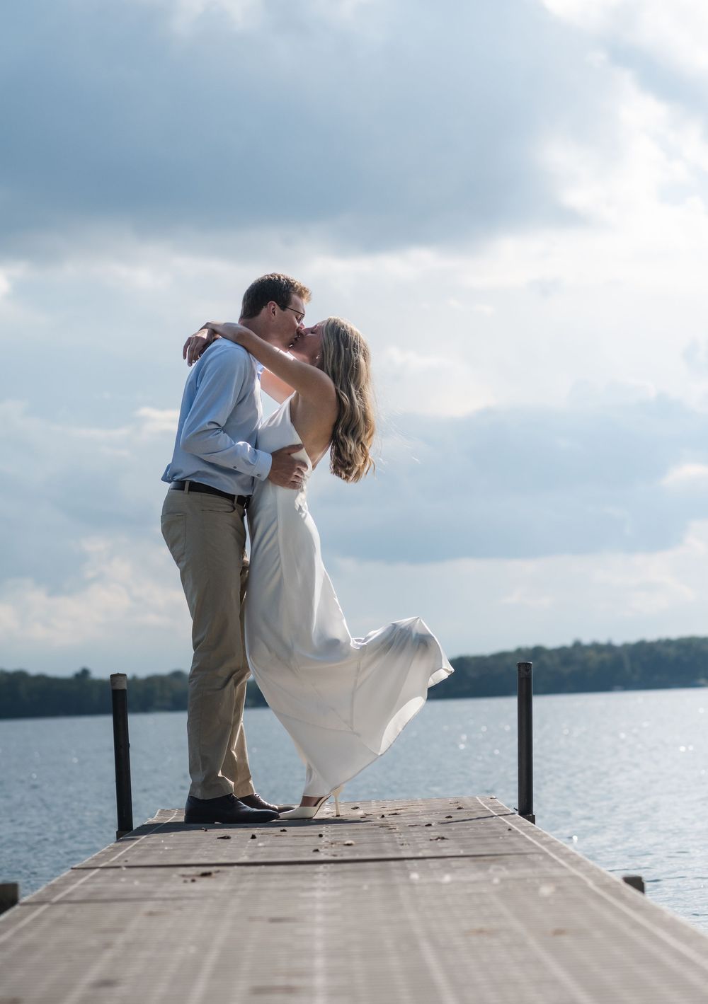 A romantic couple shares a tender moment on a wooden dock by a serene lake under cloudy skies.