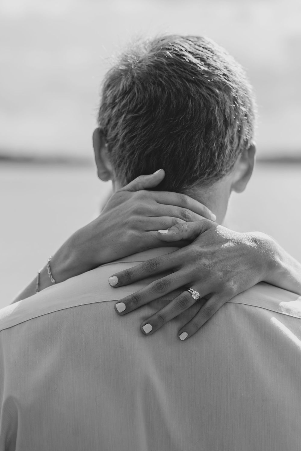 Black and white photo showing hands with an engagement ring embracing the back of someone's neck.