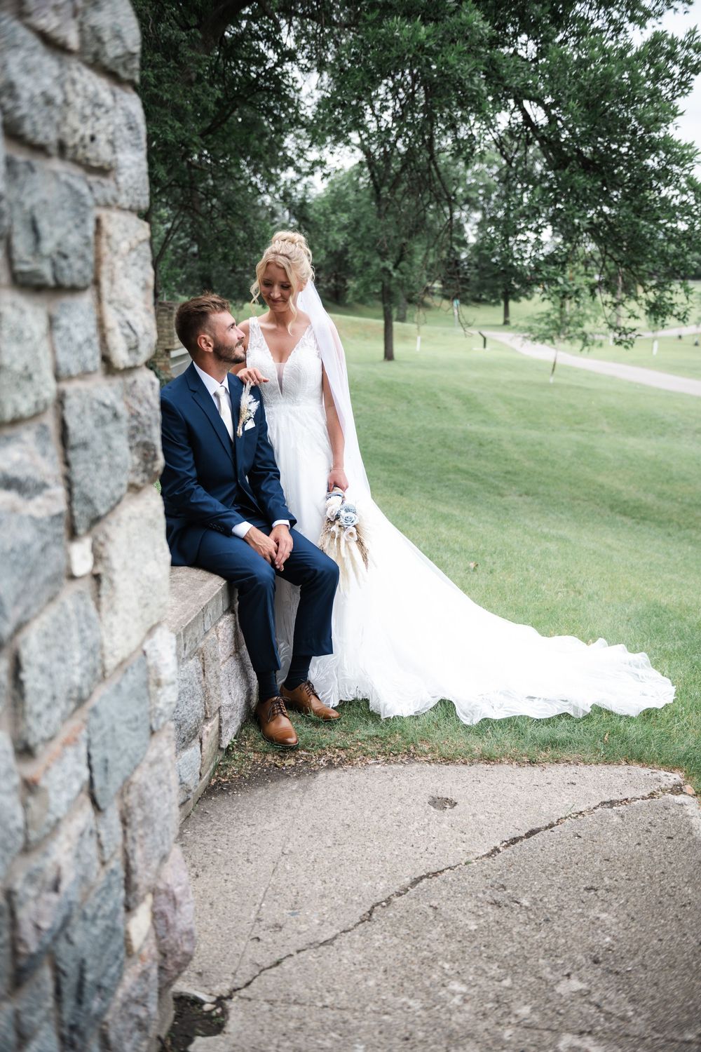 Couple in wedding attire sits on stone wall overlooking golf course.
