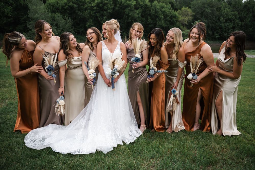 A fun bridal party in white dresses and brown jackets poses playfully in a rural outdoor setting.