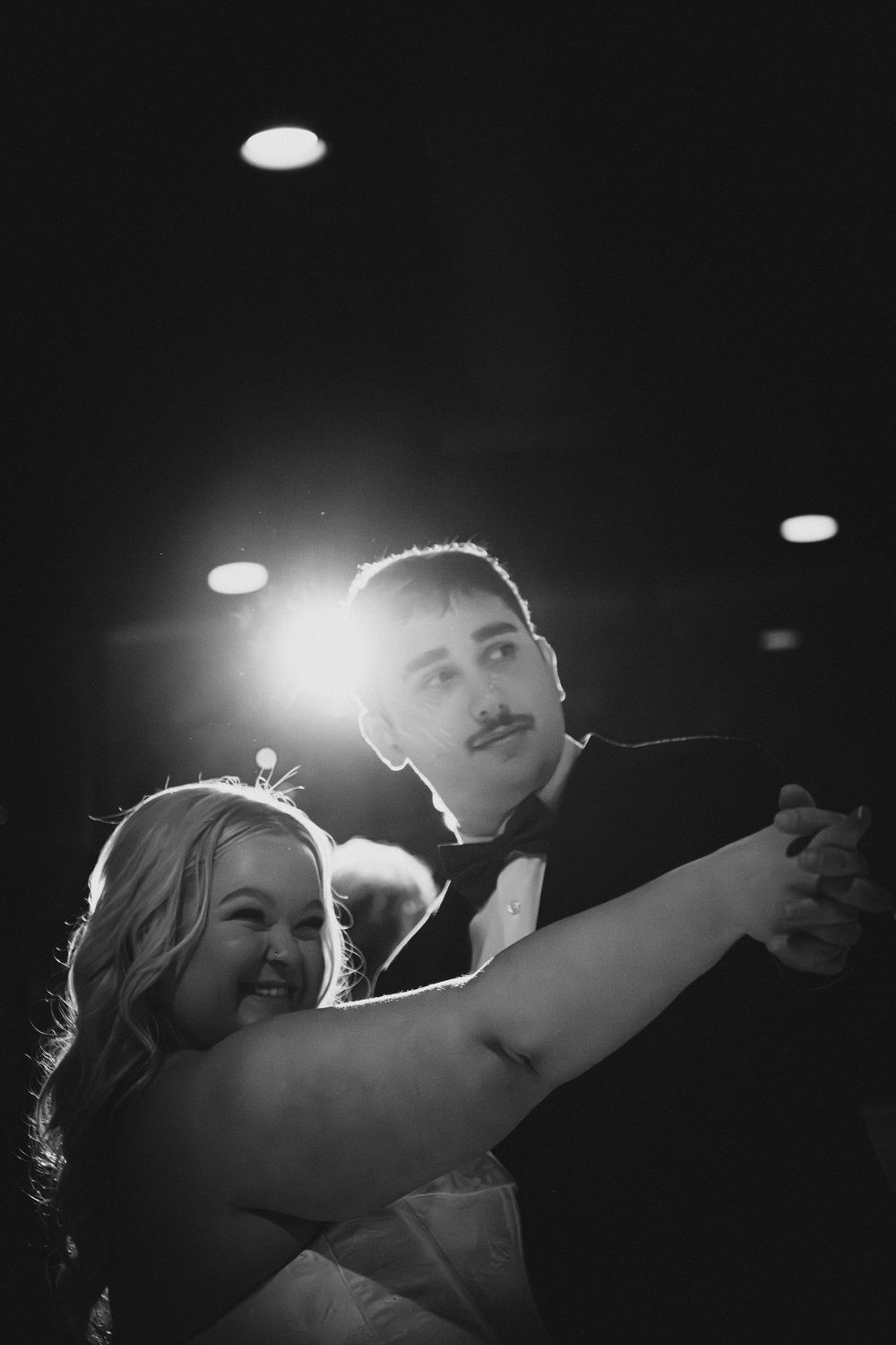 A couple dances together at their wedding reception, captured in dramatic black and white lighting.