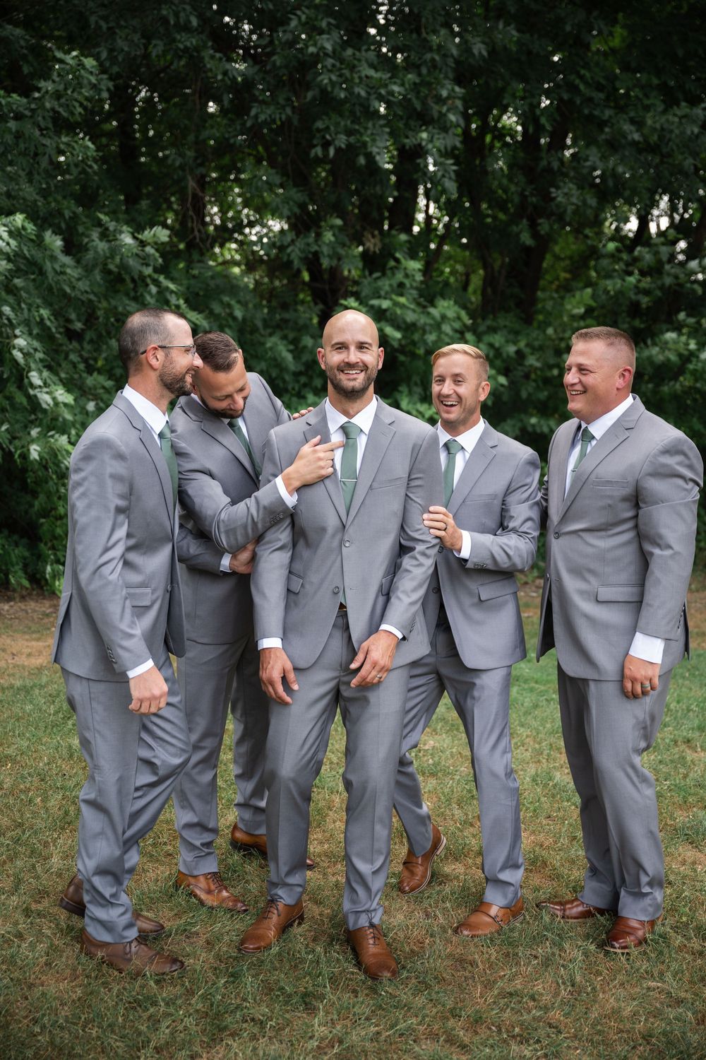 Groom and groomsmen in matching gray suits with green ties laughing together outdoors on a wedding day.