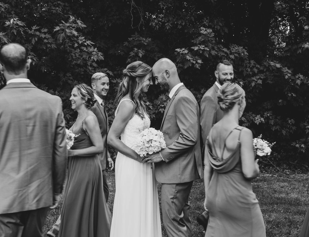 Black and white wedding party photo with bride and groom sharing intimate moment surrounded by bridesmaids and groomsmen outdoors.