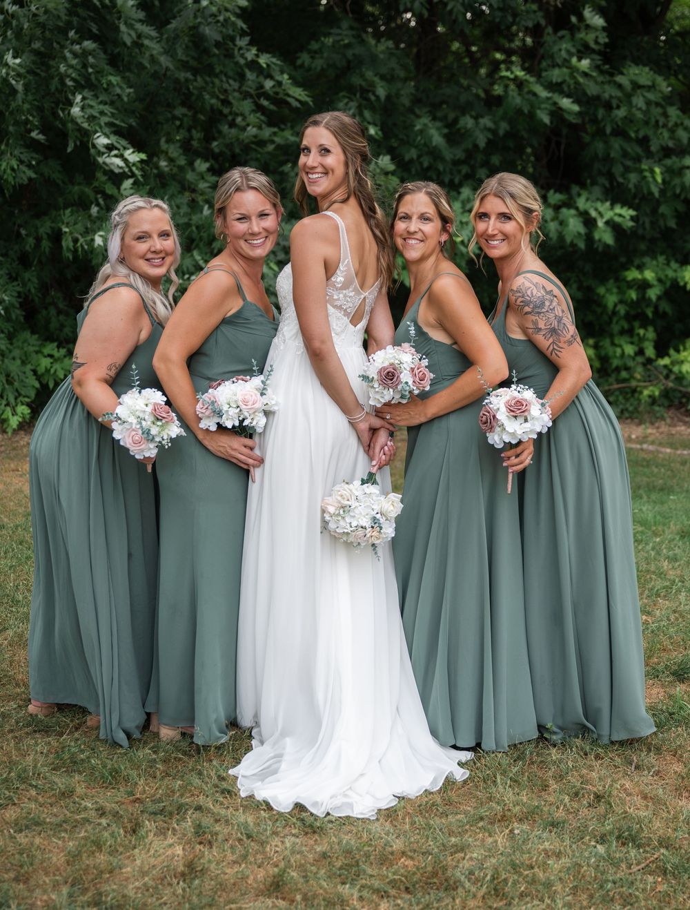 Bride in white gown poses with four bridesmaids in sage green dresses holding floral bouquets outdoors.