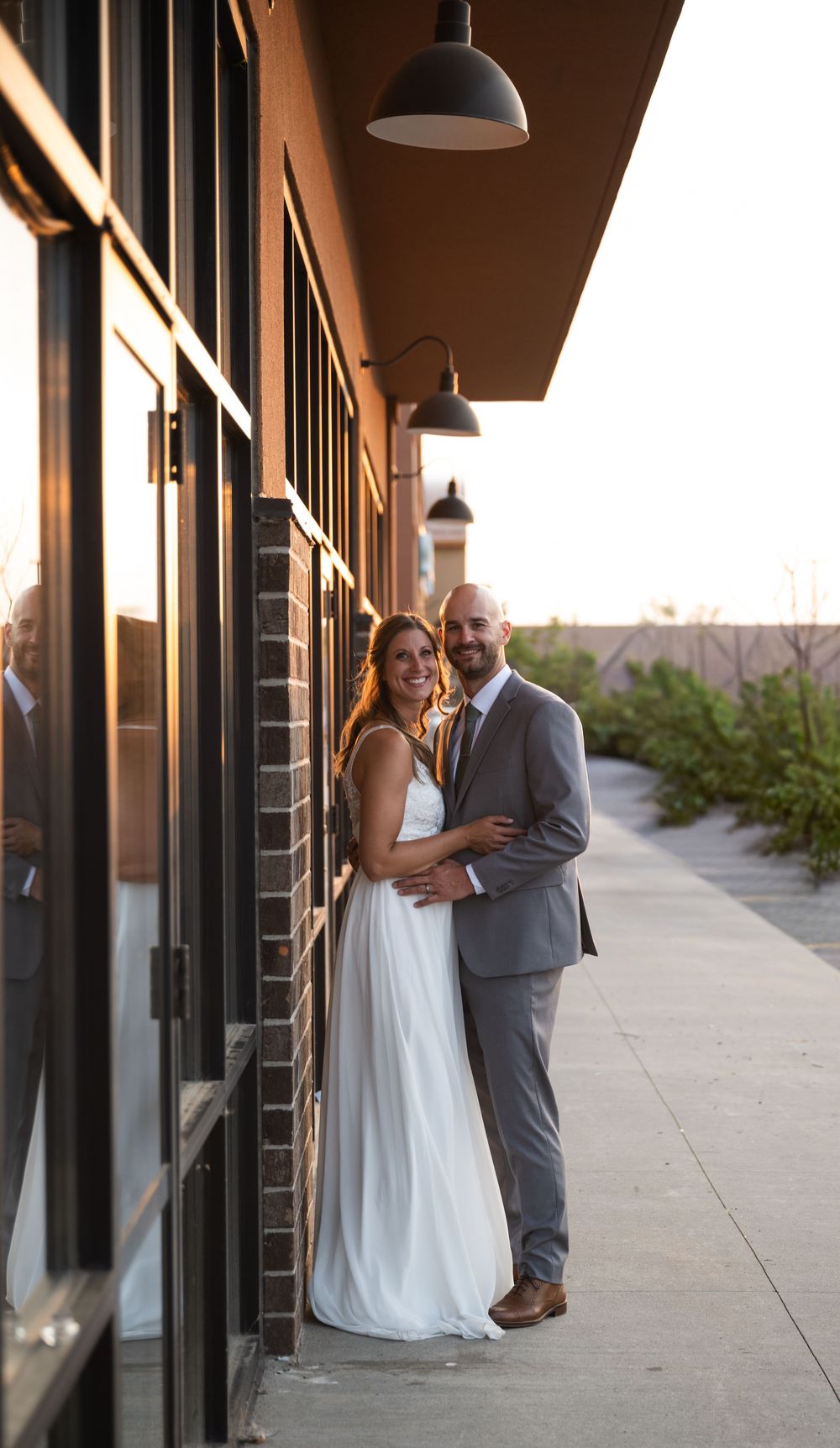 A smiling bride in a white gown and groom in a gray suit pose together outside a brick building at golden hour.