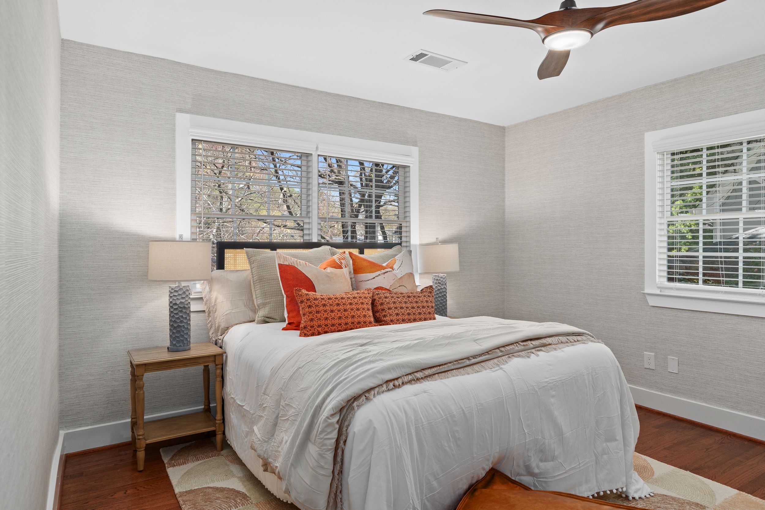 A modern bedroom with white bedding, orange accent pillows, wood floors, and a ceiling fan. Real estate photography.