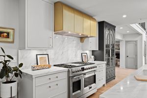 Modern white kitchen with marble backsplash, stainless steel appliances, brass upper hood, and wrap-around countertops. Real estate photo.