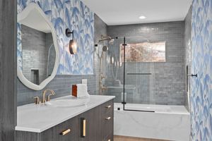 Modern bathroom with gray tile, blue patterned wallpaper, glass shower enclosure, and white vanity with circular mirror. Real estate photo.