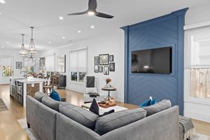 Modern open concept living room features a gray sectional sofa and blue accent wall with mounted TV. Real estate photography.
