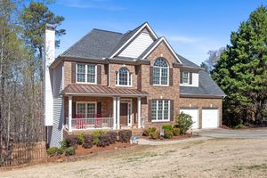 Large two-story brick suburban home with covered front porch and attached two-car garage on wooded lot. Real estate photography.