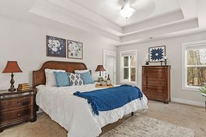 Spacious master bedroom with white bedding, blue accents, wooden furniture, and tray ceiling details. Real estate photography.