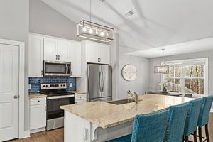 Modern white kitchen with granite countertops, stainless steel appliances, and vibrant turquoise bar stools at island. Real estate photo.