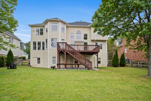 Large three-story suburban home with beige exterior and multi-level wooden deck overlooking spacious green backyard. Real estate photography