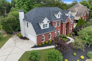 Aerial drone view of two-story colonial house with driveways and green landscaping on a suburban street. Real estate photography.