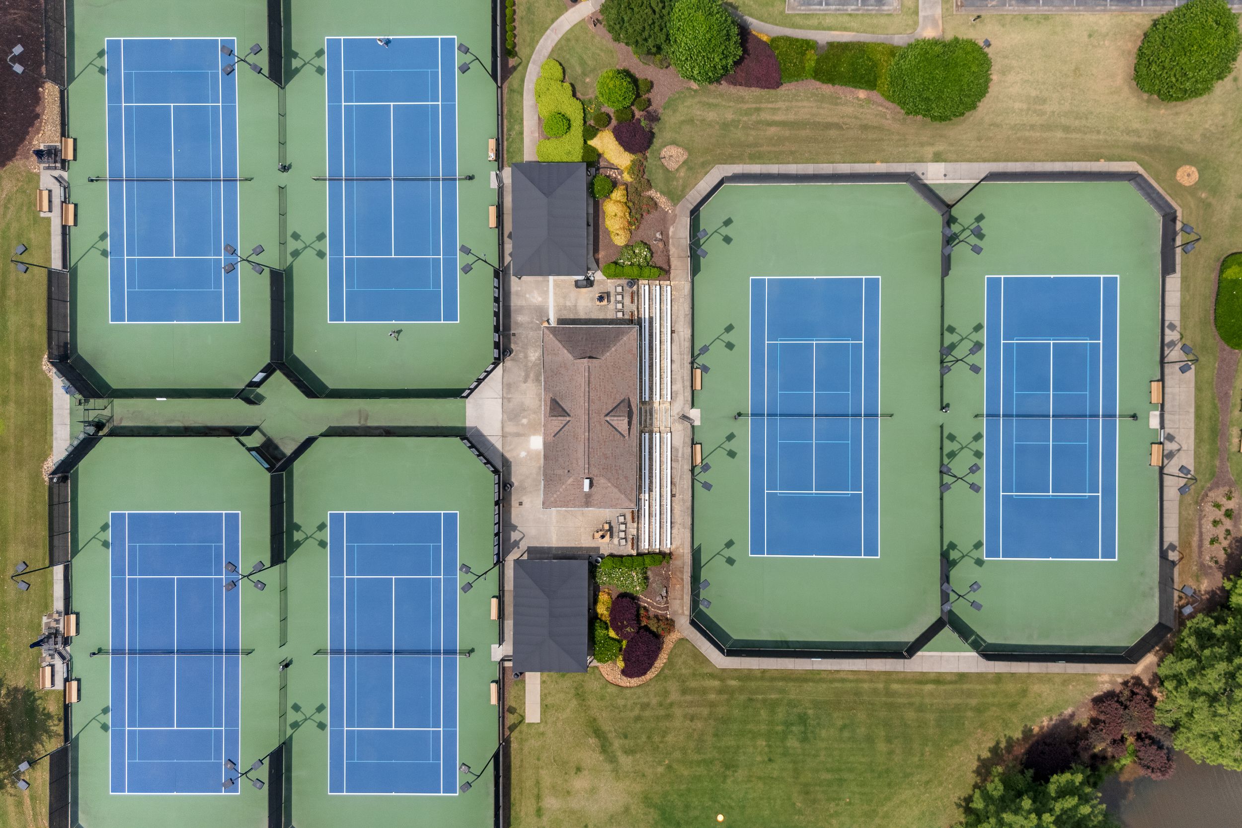 Aerial view of six blue tennis courts surrounded by green grounds and landscape features at a sports facility. Real estate photography.