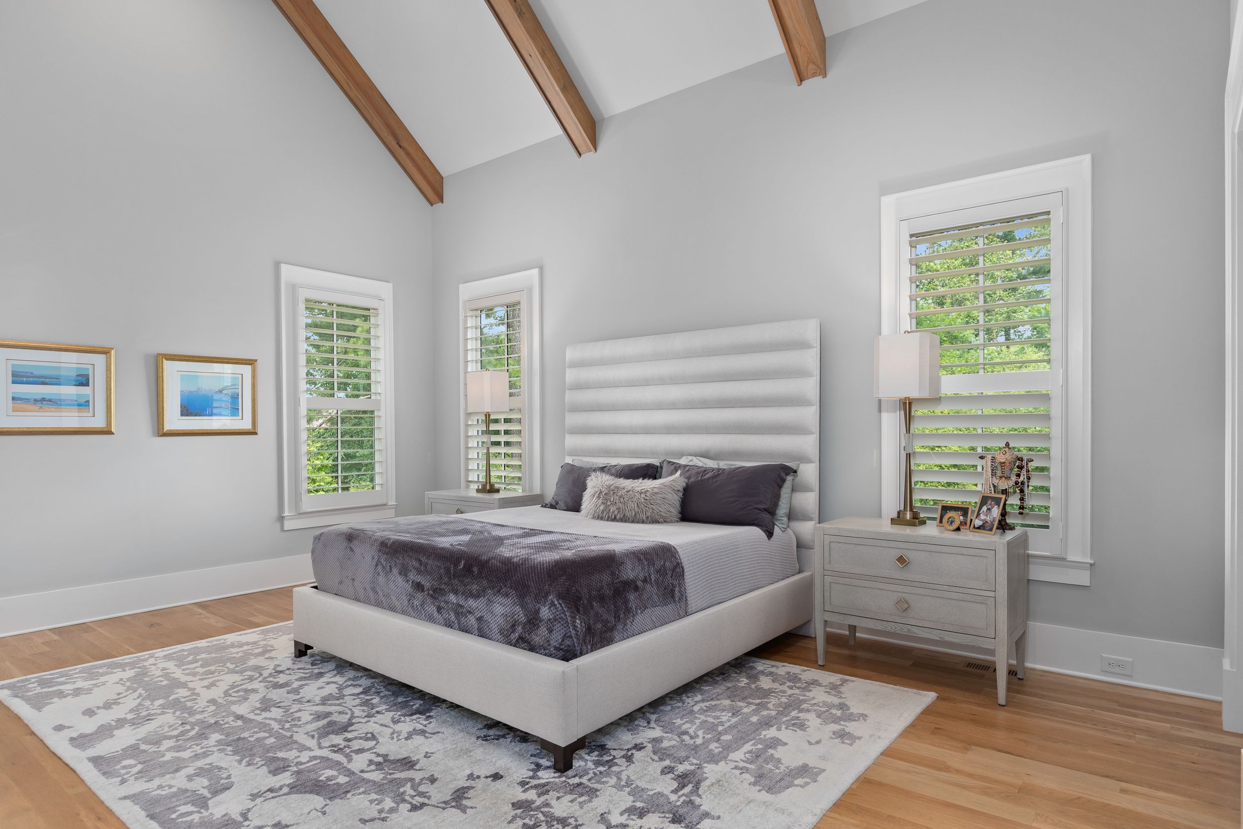 Modern bedroom with vaulted ceiling, exposed wooden beams, hardwood floors, and a gray king bed with matching decor. Real estate photography