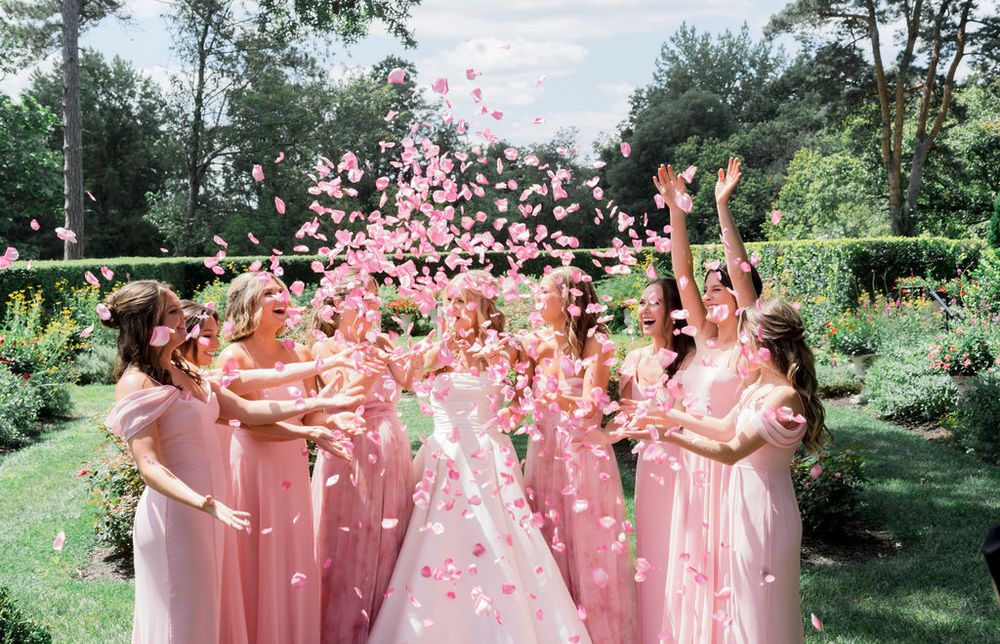 People in pink dresses stand outside in a garden setting tossing pink flower petals into the air during a celebration.