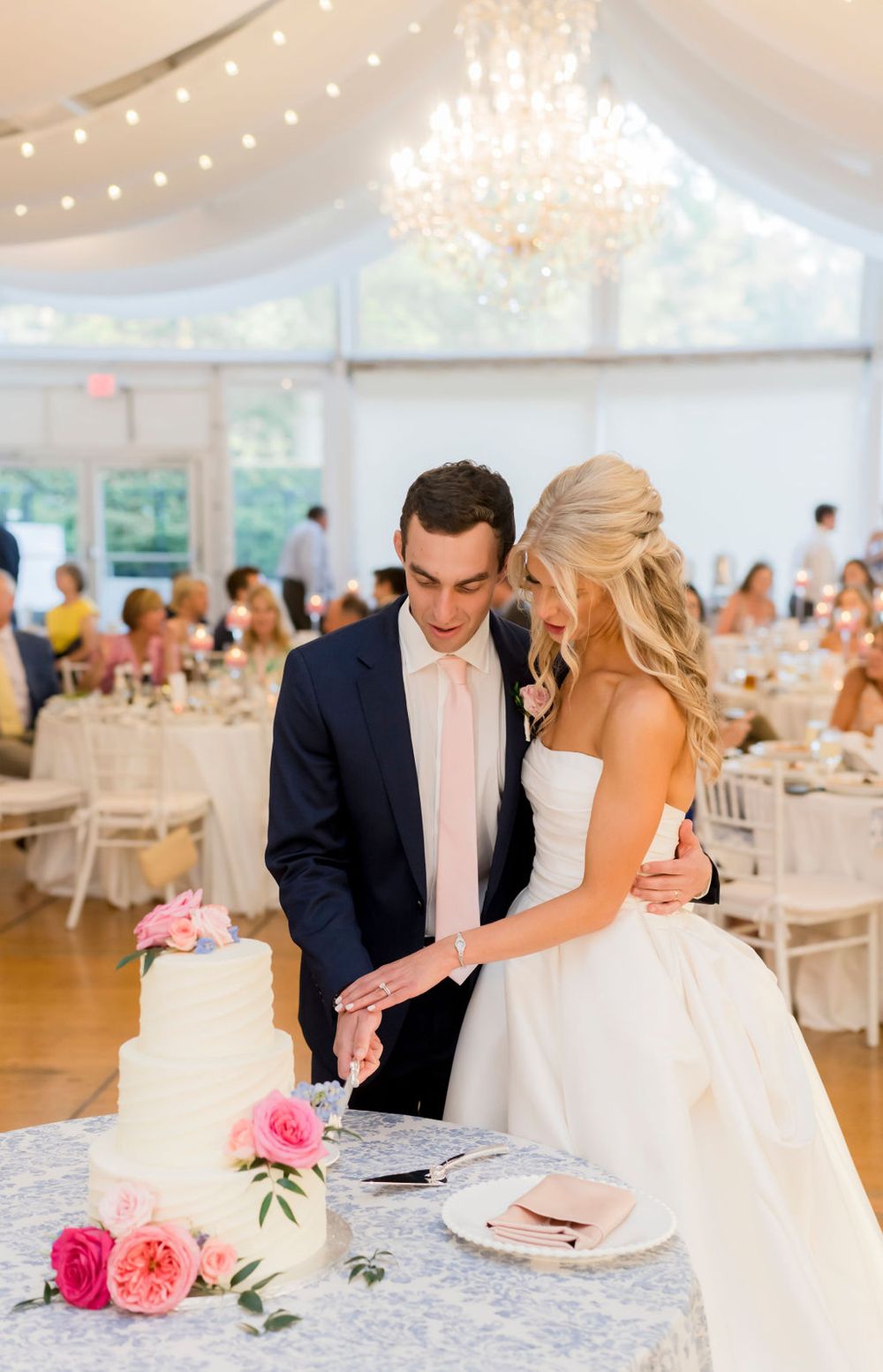 Couple cutting their elegant white wedding cake surrounded by pink flowers in a reception tent with chandelier.