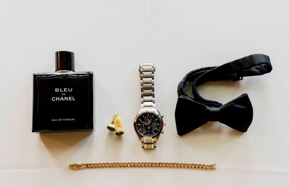 A collection of men's accessories including gold bracelet, black bow ties, wristwatch, and Chanel Bleu cologne laid on white surface.