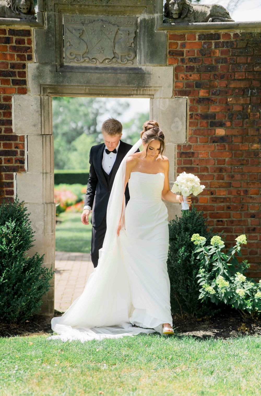Wedding couple poses under brick archways surrounded by green landscaping in an elegant outdoor setting.