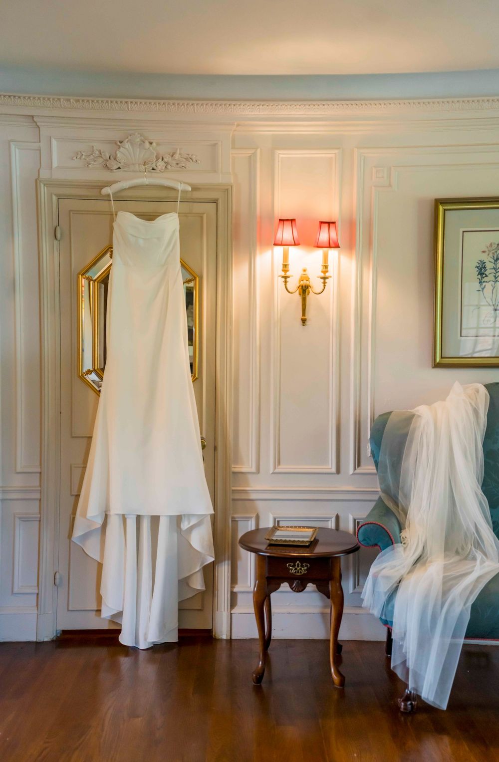 White wedding dress hanging on ornate wall with veil draped on chair and elegant sconce lighting in classic room.