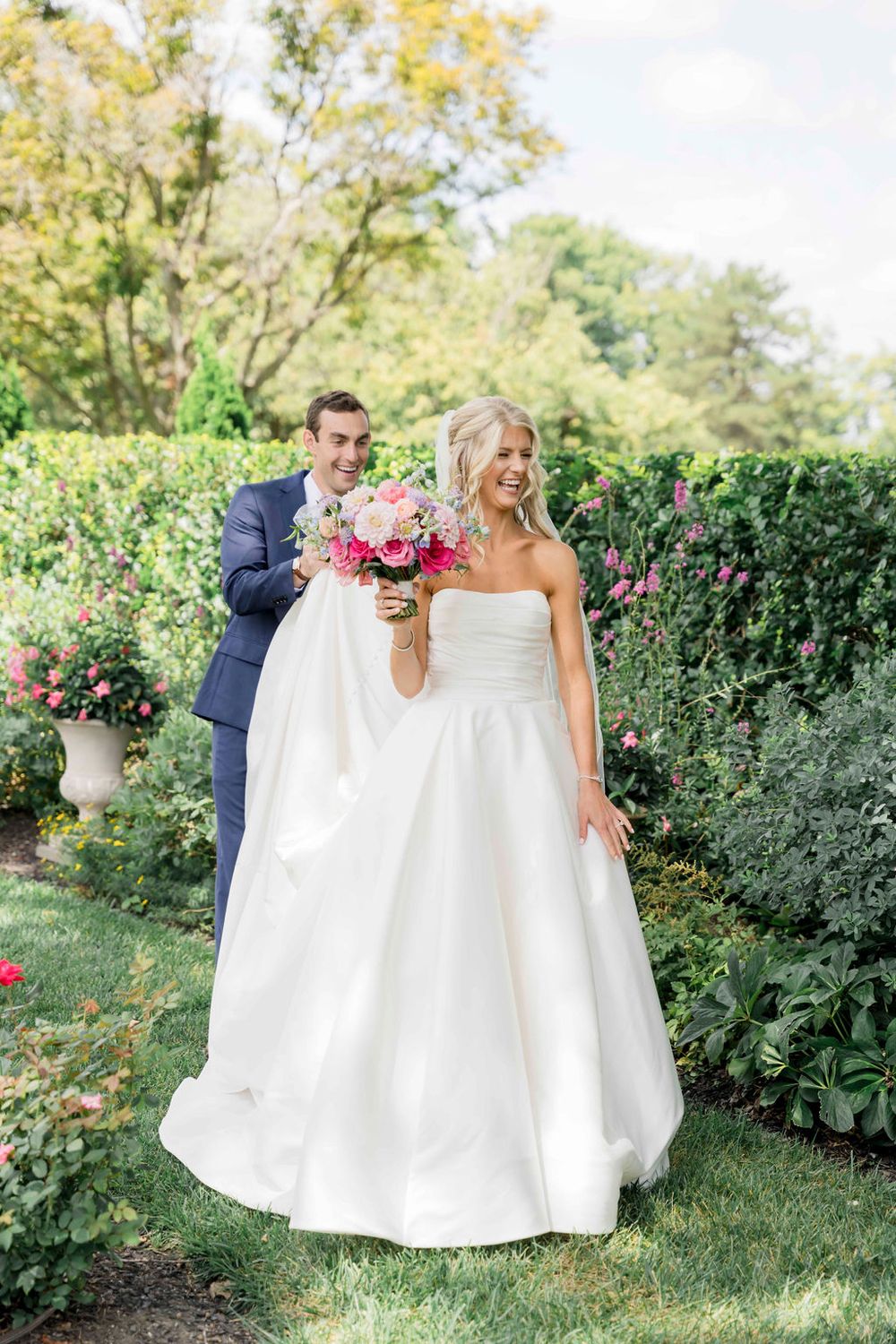 A couple celebrates in a lush garden setting with elegant wedding attire and pink floral bouquet on a sunny day.