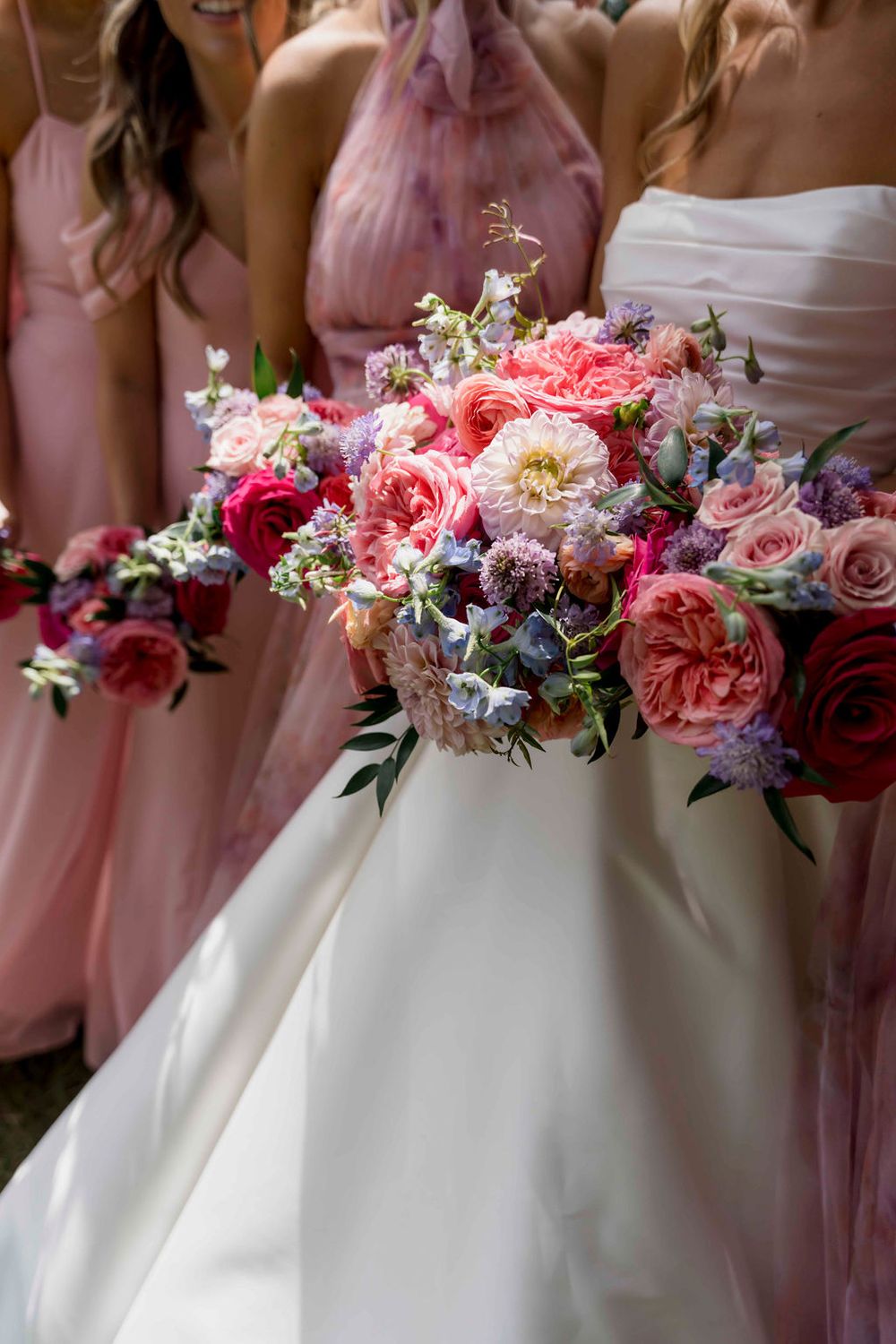 Pink and coral rose bouquets sit beautifully against white and blush wedding dresses in an elegant bridal party lineup.
