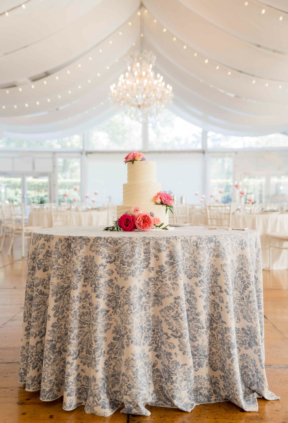 Wedding reception tent with white draping, crystal chandelier and floral cake display on patterned tablecloth.