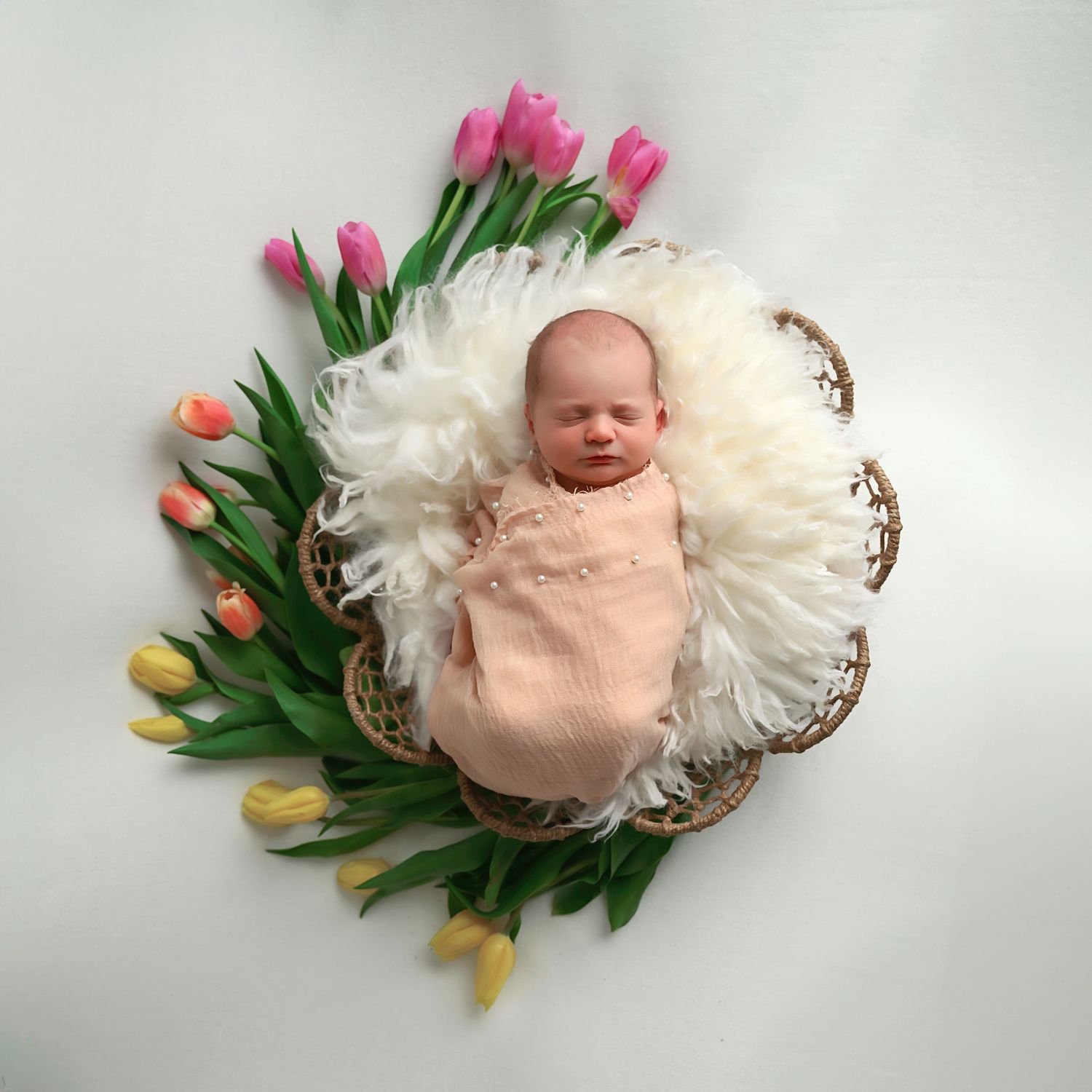 Newborn baby wrapped in peach blanket lies in basket with white fur, surrounded by colorful spring tulips.