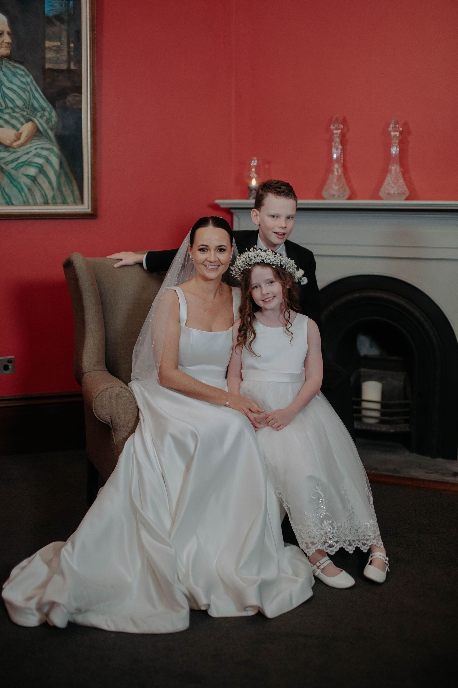 A bride in white gown sits on brown chair with two children in formal attire beside fireplace with red walls.