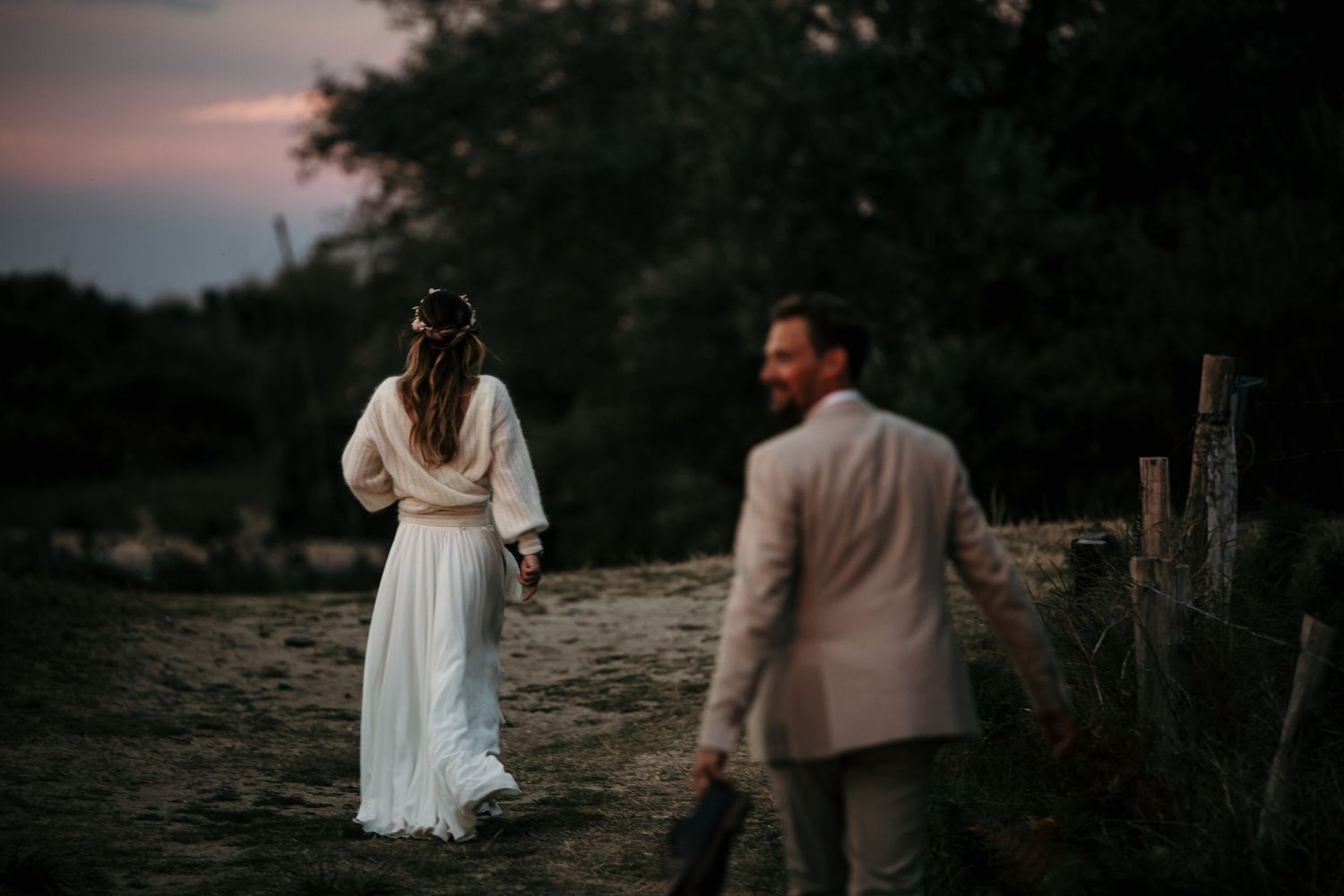 Couple in wedding attire stand in a moody twilight setting near a wooden fence.