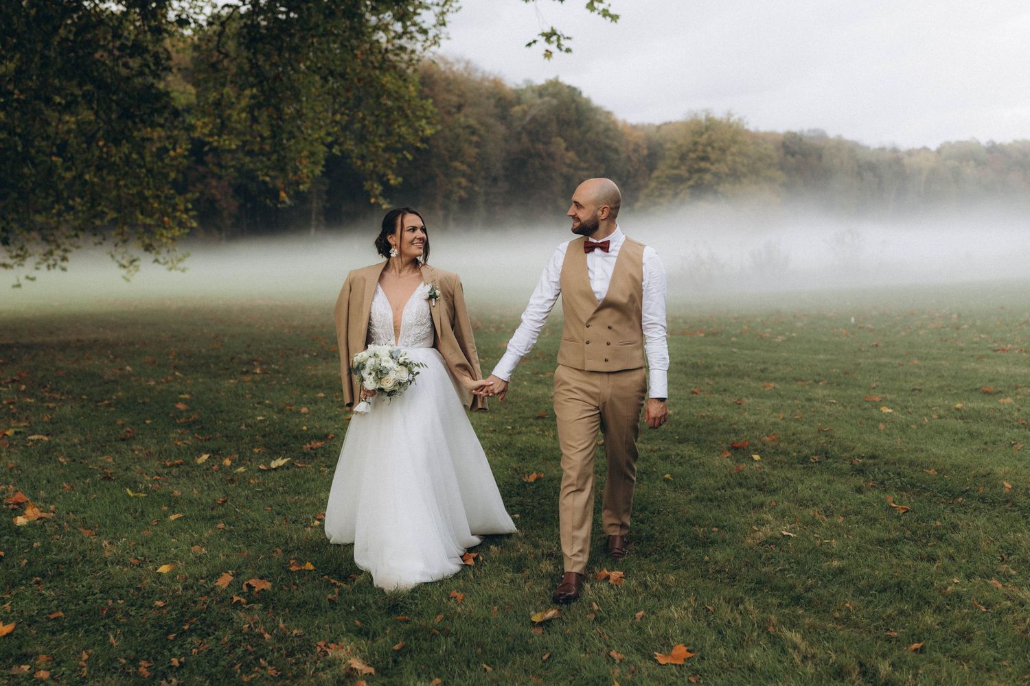 A couple in wedding attire walks hand in hand through a misty autumn field during a romantic outdoor ceremony.