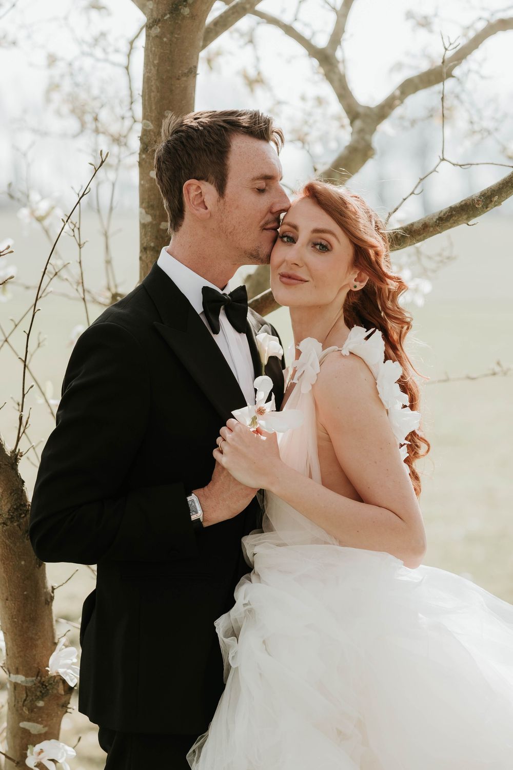 A romantic couple in editorial wedding attire embrace outdoors beneath winter trees in soft dreamy light at Norwood park.