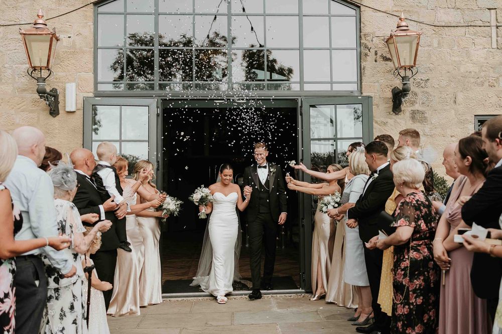 A wedding party celebrates with confetti outside a rustic stone venue of Wharfedale grange with lanterns and large glass windows.