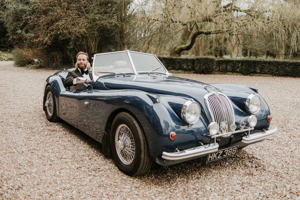A classic vintage Jaguar XK140 roadster in metallic blue arriving at Wootton hall with groom driving wearing black tie.